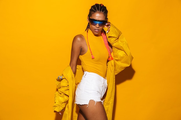 Bild einer wunderschönen afroamerikanischen Frau mit Sonnenbrille und Windjacke, die isoliert über gelber Wand posiert?