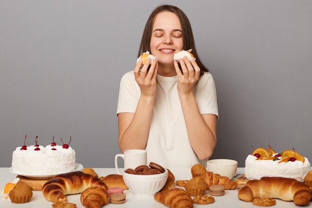 Bild einer zufriedenen, fröhlichen, entzückenden Frau mit braunen Haaren, die ein weißes T-Shirt trägt und am Tisch sitzt, mit Desserts, die ein angenehmes Aroma nach Kuchen riechen, isoliert auf grauem Hintergrund