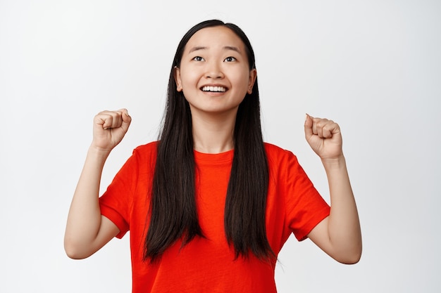 Bild eines asiatischen Mädchens, das lächelt, hoffnungsvoll aufschaut und singt, jubelt als Sportspiel, stehend im roten T-Shirt auf Weiß.