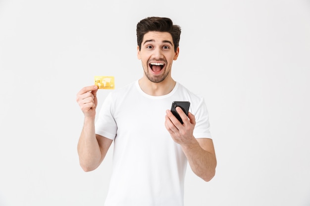 Bild eines aufgeregten, glücklichen jungen Mannes, der isoliert über der weißen Wand posiert und das Mobiltelefon mit Kreditkarte verwendet.