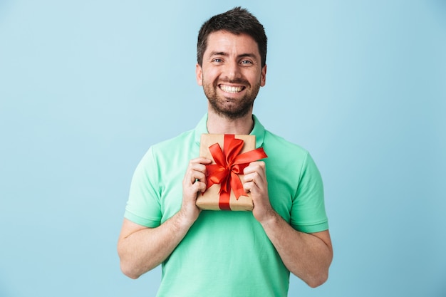 Bild eines aufgeregten jungen, gutaussehenden bärtigen Mannes, der isoliert über der blauen Wand posiert, die eine Geschenkbox hält.