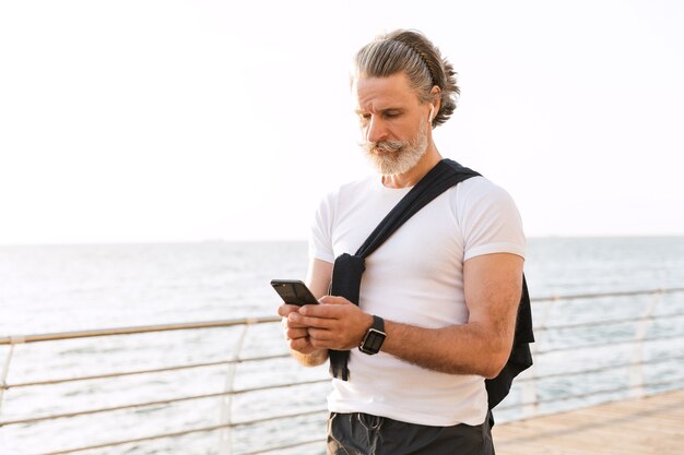 Bild eines denkenden alten Mannes in Sportkleidung mit Ohrstöpsel und Handy beim Training an der Promenade am Morgen