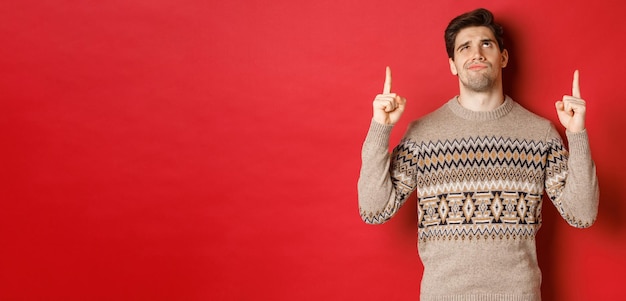 Bild eines enttäuschten und skeptischen gutaussehenden Mannes, der einen Weihnachtspullover trägt und die Stirn runzelt, während er auf etwas Schlechtes blickt und mit dem Finger nach oben zeigt, der über rotem Hintergrund steht