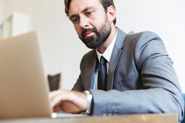 Bild eines ernsthaften bärtigen Geschäftsmannes, der im Büro sitzt und einen Laptop verwendet, der drinnen arbeitet.