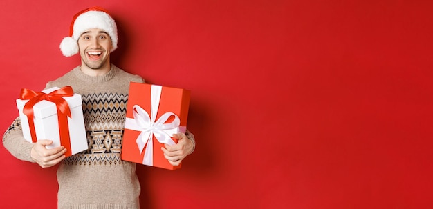 Bild eines fröhlichen, attraktiven Kerls, der Weihnachtsgeschenke hält, in Weihnachtsmütze und Winterpullover steht, glücklich lächelt und über rotem Hintergrund steht.
