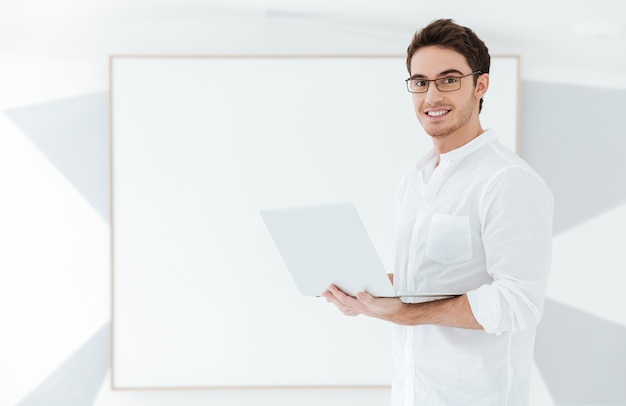 Bild eines fröhlichen Mannes mit Brille und gekleidet in weißem Hemd mit Laptop-Computer in der Nähe von Big Board. Schauen Sie in die Kamera.