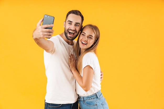 Bild eines fröhlichen Paares in Denim-Kleidung, das lächelt, während es ein Selfie-Foto auf dem Handy macht, isoliert über gelber Wand