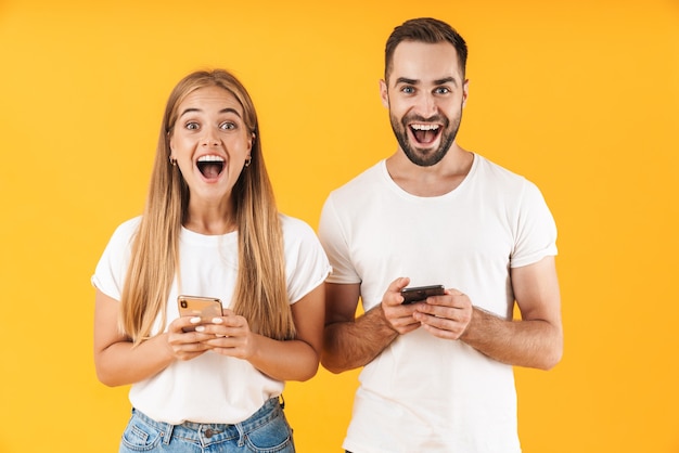 Bild eines fröhlichen Paares Mann und Frau, die lächeln, während sie Smartphones zusammen verwenden, isoliert über gelber Wand?