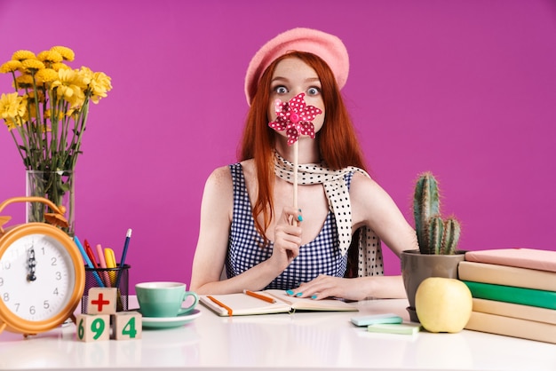 Bild eines fröhlichen Teenager-Mädchens, das Papierblume auf einem Stock hält, während es mit Schulheften am Schreibtisch studiert, isoliert über der violetten Wand?