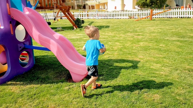 Bild eines glücklich lachenden fröhlichen Jungen, der Fußballball in den Händen hält und auf dem Kinderspielplatz im Park läuft