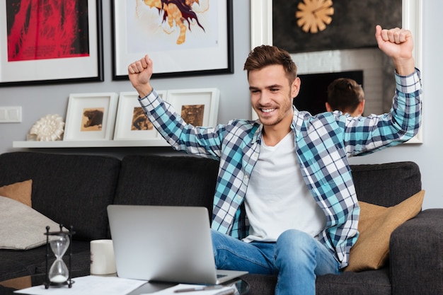 Bild eines glücklichen Borstenmannes, der in einem Hemd in einem Käfigdruck auf dem Sofa zu Hause sitzt und einen Laptop verwendet