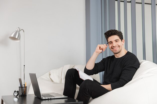 Bild eines glücklichen gutaussehenden jungen Mannes zu Hause drinnen unter Verwendung des Laptops.