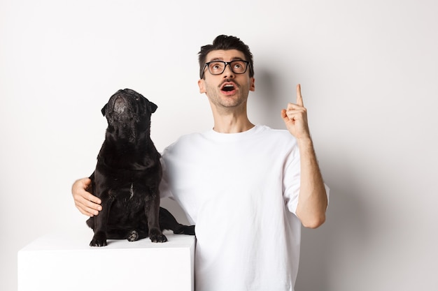 Bild eines glücklichen jungen Mannes und eines glücklichen Hundes, die beide zur Promo aufblicken, der Besitzer zeigt mit dem Finger nach oben, der Mops starrt auf den Klo, der auf weißem Hintergrund steht