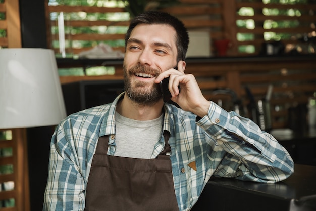 Bild eines glücklichen Kellnerjungen mit Schürze, der an der Bar sitzt und Handy benutzt, während er im Café oder Kaffeehaus im Freien arbeitet?