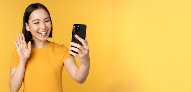 Bild eines glücklichen, schönen asiatischen Mädchens, das Video-Chats auf einer Smartphone-Anwendung spricht, die vor gelbem Hintergrund steht Kopieren Sie Platz