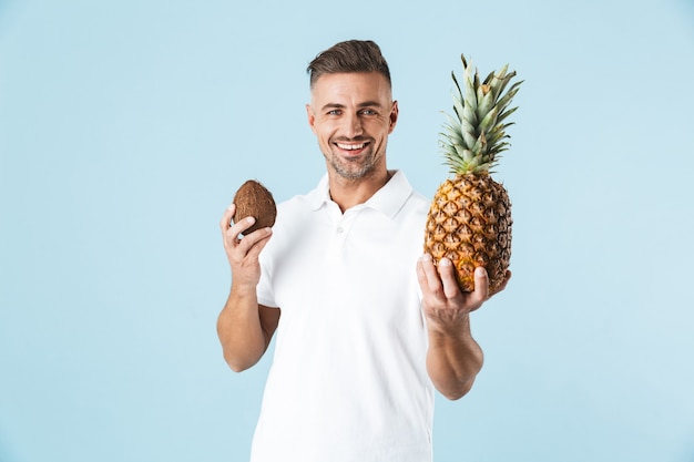 Bild eines gutaussehenden aufgeregten glücklichen erwachsenen Mannes, der über der blauen Wand hält, die Kokosnuss und Ananas hält.