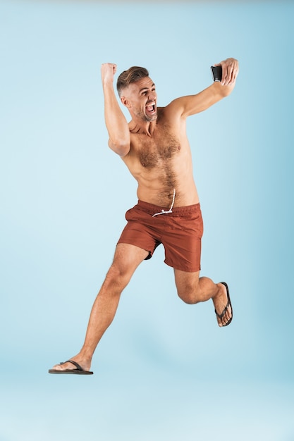 Bild eines gutaussehenden aufgeregten glücklichen erwachsenen Mannes in der Badebekleidung, die über das Springen der blauen Wand aufwirft, machen Selfie durch Handy.