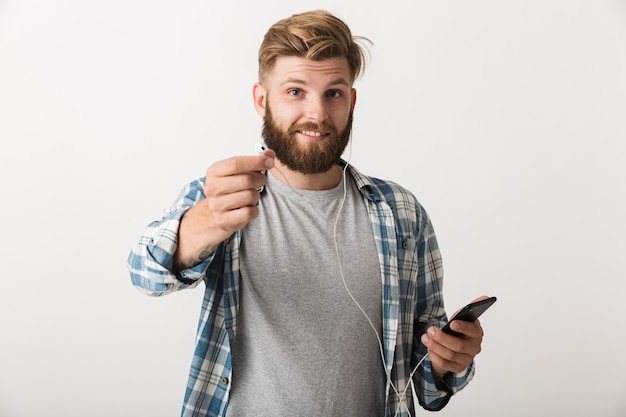 Bild eines gutaussehenden jungen bärtigen Mannes, der lokalisiert über weißem Wandhintergrund unter Verwendung der hörenden Musik des Mobiltelefons steht.