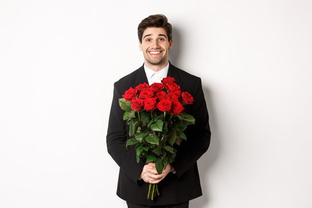 Bild eines gutaussehenden Mannes im schwarzen Anzug, der einen Rosenstrauß hält und lächelt und vor weißem Hintergrund steht
