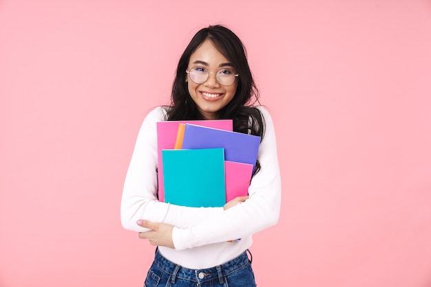 Bild eines jungen brünetten asiatischen Studentenmädchens mit Brille, das Papierordner isoliert auf Rosa hält