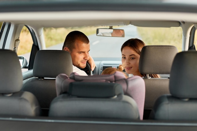 Bild eines jungen erwachsenen Mannes und einer jungen Frau mit kleinem Kind, die im Auto fahren, Babytochter, die im Autositz sitzt, Vater und Mutter, die auf ihr Kind zurückblicken