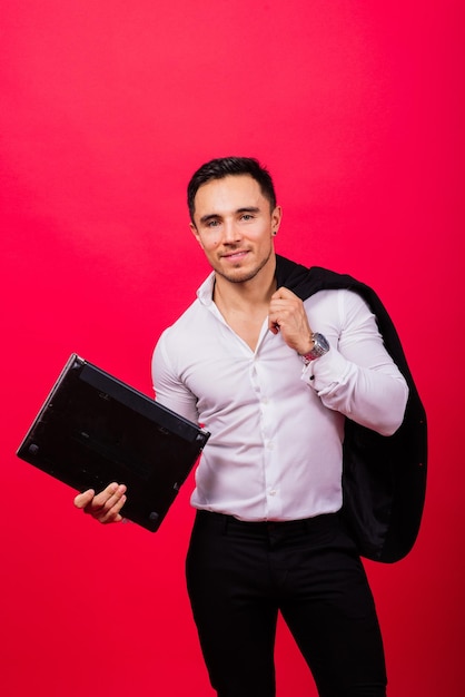 Bild eines jungen fröhlichen Geschäftsmannes in der Uhr, der Laptop isoliert auf rotem Hintergrund hält und verwendet