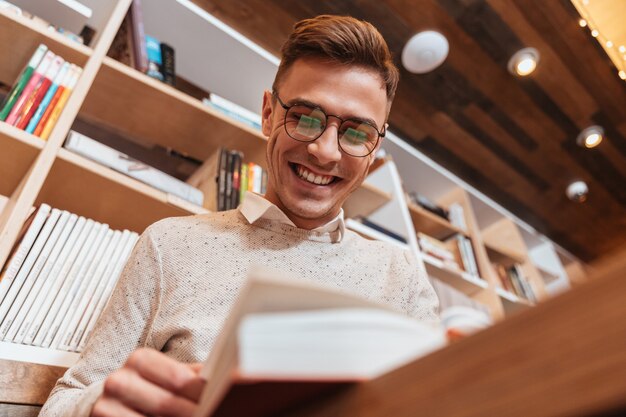 Bild eines jungen glücklichen Mannes mit Brille im Hemd, der im Café sitzt, während er ein Buch liest. Schauen Sie sich das Buch an.