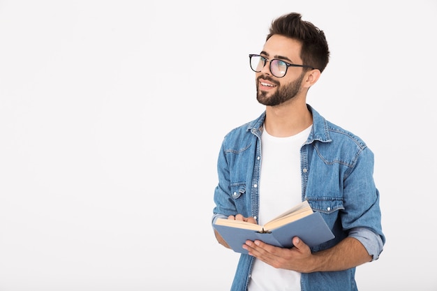 Bild eines jungen intelligenten Mannes, der eine Brille trägt, ein Buch hält und den Kopierraum isoliert über der weißen Wand beiseite schaut