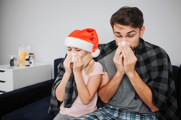 Bild eines jungen Mannes mit Tochter, die auf dem Sofa sitzt und niest. Sie haben sich erkältet. Grippe und Krankheit. Das neue Weihnachtsjahr feiern. Roter Hut des Kopfes des Mädchens. Festlicher Monat