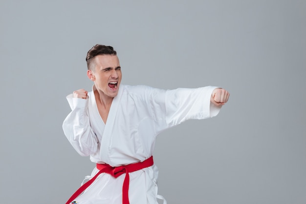 Bild eines jungen Sportlers im Kimono, der Karate übt, während er auf grauem Hintergrund posiert und schreit. Beiseite schauen.