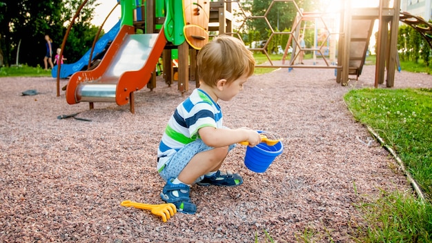 Bild eines kleinen Jungen, der auf dem Spielplatz sitzt und Sand mit einem kleinen Plastikspaten in einem bunten Eimer reinigt. Kind gräbt und baut im Park