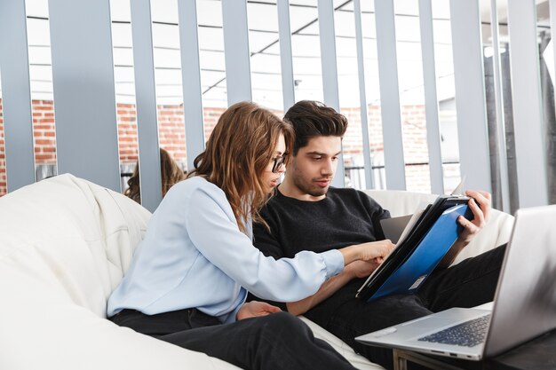 Bild eines konzentrierten jungen liebenden Paares zu Hause drinnen unter Verwendung der Laptop-Computerarbeit mit Dokumenten.