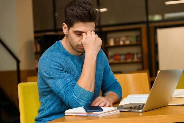 Bild eines konzentrierten Mannes eines jungen Studenten mit Kopfschmerzen in der Bibliothek, die Hausaufgaben macht, die lesen und Laptop-Computer verwenden.