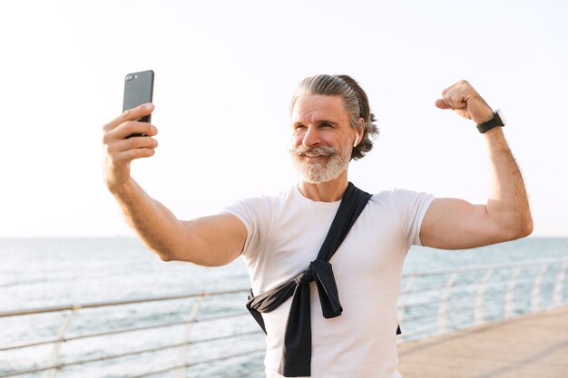 Bild eines lächelnden alten Mannes in Sportkleidung, der seinen Bizeps zeigt, während er morgens ein Selfie-Foto auf dem Handy an der Promenade macht