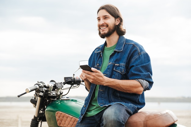 Bild eines lächelnden glücklichen glücklichen Mannradfahrers auf seinem Fahrrad draußen an einem Strand mit Handy-Chat.