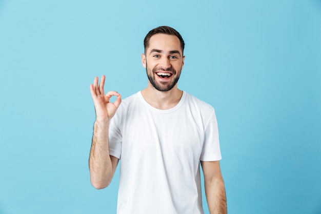 Bild eines lächelnden jungen, glücklichen bärtigen Mannes, der isoliert über der blauen Wand posiert und eine gute Geste zeigt. Sommer-Konzept.