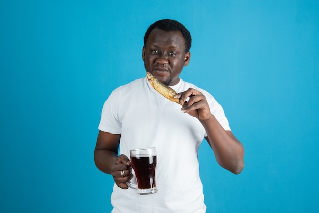 Bild eines Mannes im weißen T-Shirt, der getrockneten Fisch mit einem Glas Wein gegen die blaue Wand hält