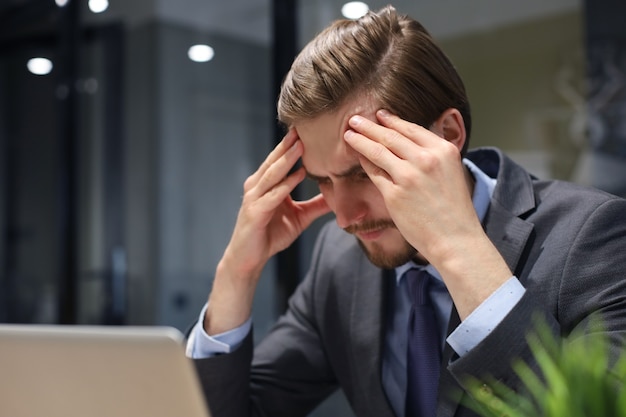 Bild eines müden jungen Geschäftsmannes im Anzug, der das Gesicht in den Händen hält, während er im Büro sitzt.