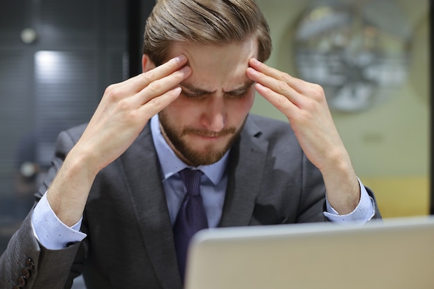 Bild eines müden jungen Geschäftsmannes im Anzug, der das Gesicht in den Händen hält, während er im Büro sitzt