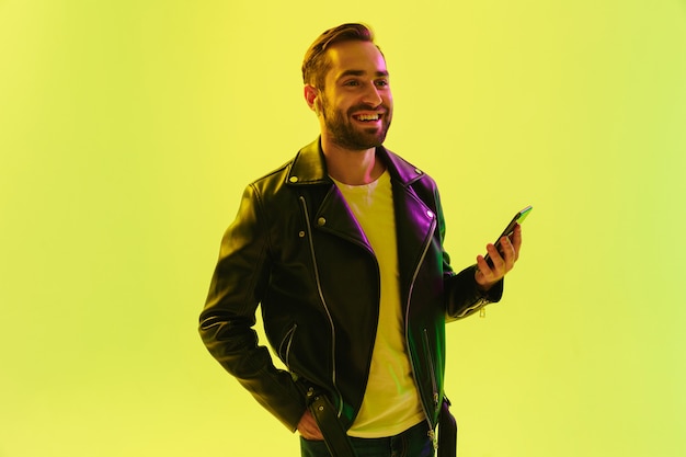 Bild eines optimistisch erfreuten, fröhlichen, gutaussehenden jungen, stilvollen Mannes, der über hellgrüner Wand mit LED-Neonlichtern in Lederjacke mit Mobiltelefon isoliert ist.