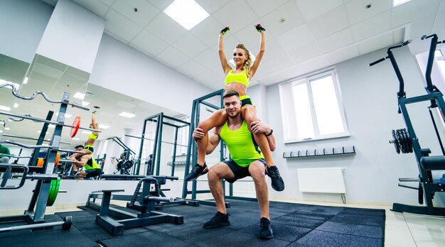 Bild eines persönlichen Fitnesstrainers und einer Kundin im Fitnessstudio, die vor der Kamera posieren Gesundes Leben und Fitnesskonzept Mädchen sitzt in Siegerpose auf der Schulter des Mannes