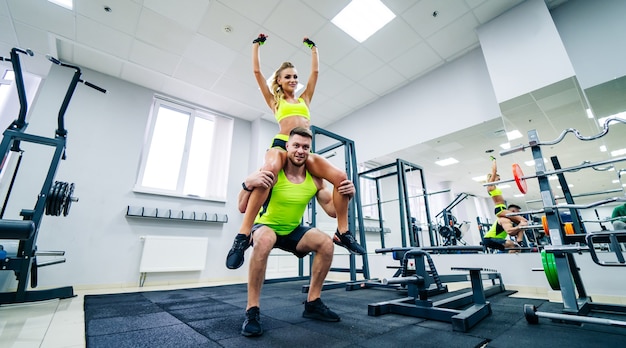 Bild eines persönlichen Fitnesstrainers und einer Kundin im Fitnessstudio, die vor der Kamera posiert. Gesundes Leben und Fitnesskonzept. Mädchen sitzt auf der Schulter des Mannes in Siegerpose.