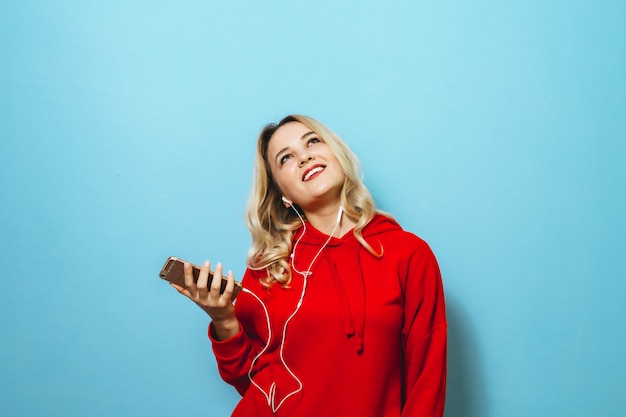 Bild eines schönen blonden aufgeregten Mädchens, das Musik in den Kopfhörern hört und über blaue Wand tanzt