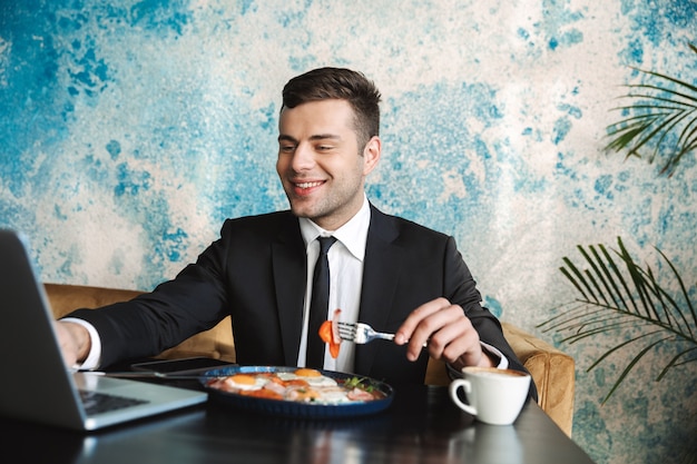 Bild eines schönen glücklichen jungen Geschäftsmannes, der im Café unter Verwendung des Laptops sitzt, haben ein Frühstück oder Abendessen essen.