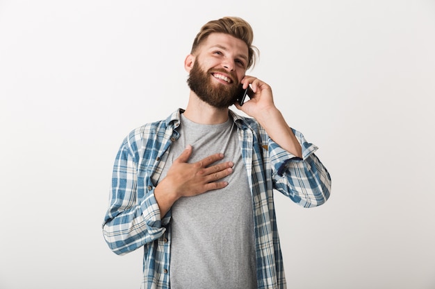 Bild eines schönen jungen bärtigen Mannes, der lokalisiert über weißem Wandhintergrund steht, der durch Handy spricht.