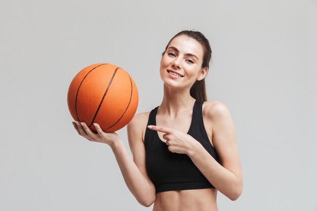 Bild eines schönen jungen sporteignungsfrauen-basketballspielers, der mit basketball lokalisiert über grauer wand aufwirft.