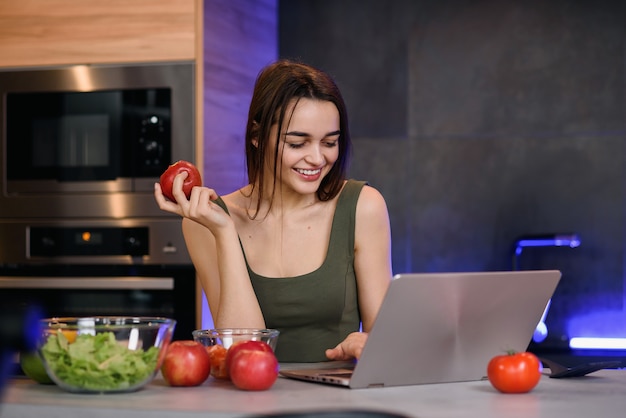 Bild eines schönen jungen Studentenmädchens mit Apfel, der drinnen unter Verwendung des Laptops sitzt.