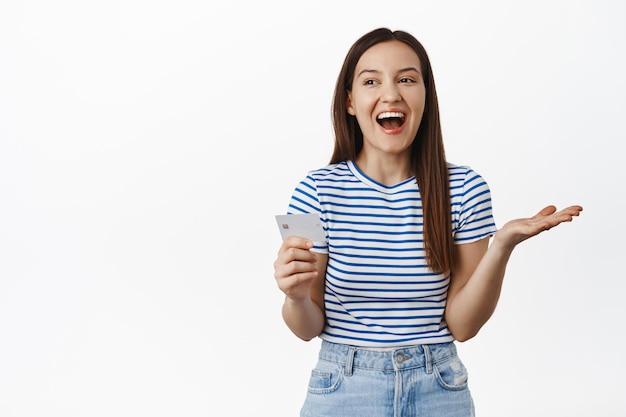 Bild eines schönen Mädchens, das lacht und lächelt, eine Kreditkarte in der Hand hält, in Freizeitkleidung steht, gestreiftes Sommer-T-Shirt, isoliert auf weißem Hintergrund.