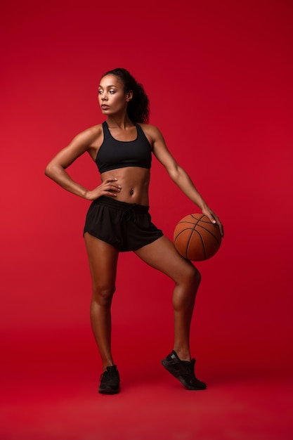Bild eines starken jungen afrikanischen Sportfitnessfrauenbasketballspielers, der lokalisiert über der roten Wandwand hält, die Ball hält.