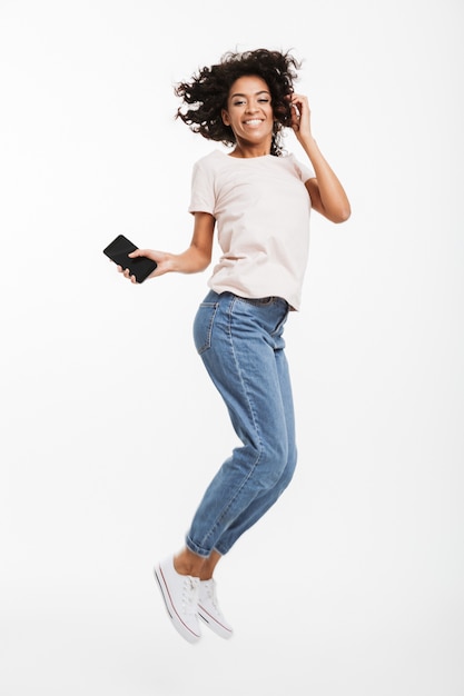 Bild in voller Länge der glücklichen Afroamerikanerfrau mit braunem lockigem Haar, das Jeans und T-Shirt trägt, die mit Smartphone in der Hand schweben, lokalisiert über weißer Wand